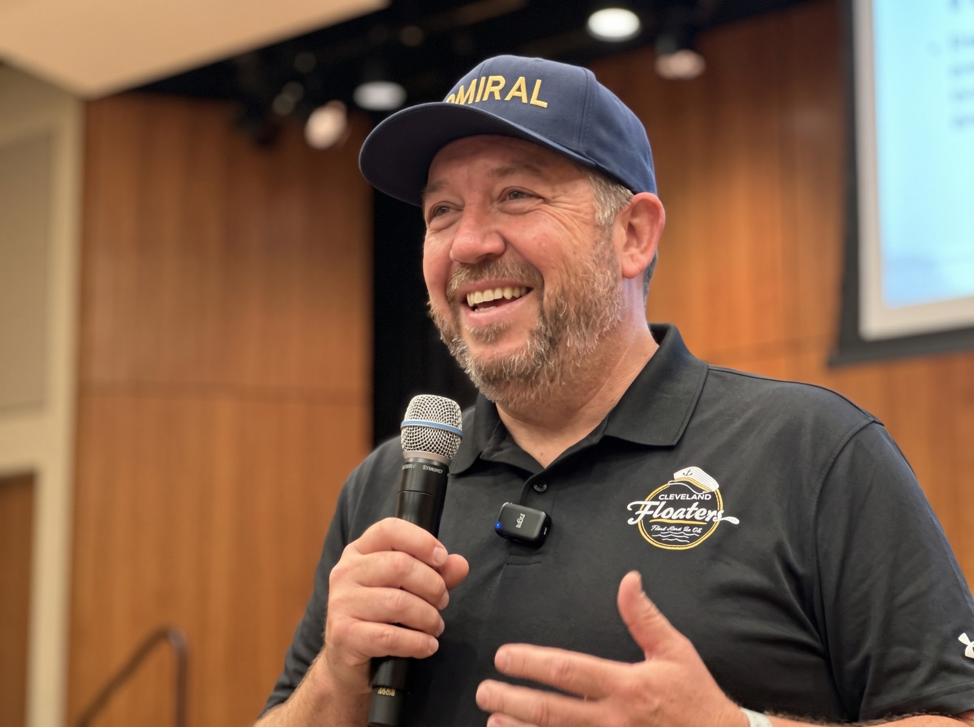 Admiral Jim in a navy ADMIRAL cap and Cleveland Floaters polo, smiling and speaking into a microphone